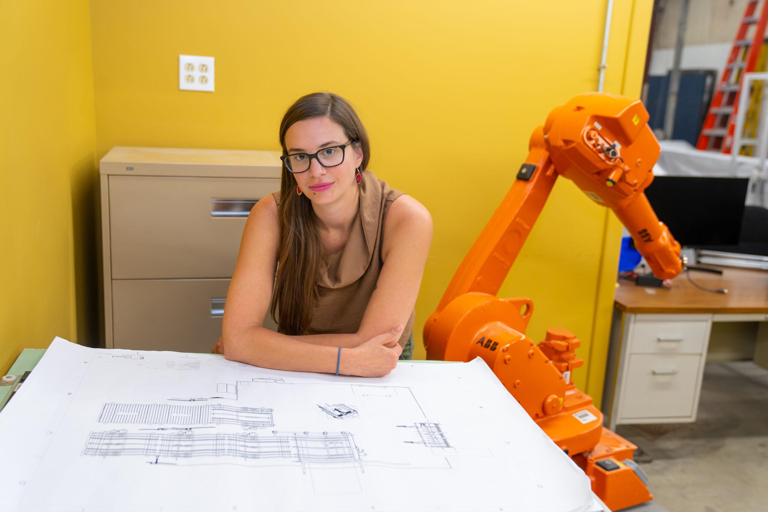Rigorous COO showing manufacturing plant blueprint with robot behind her.